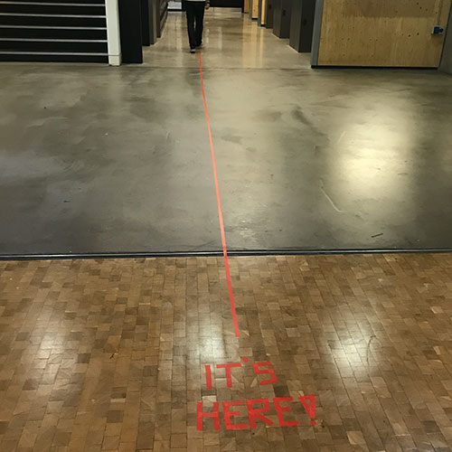The words It's here in red taped on the floor with a red line leading out of frame